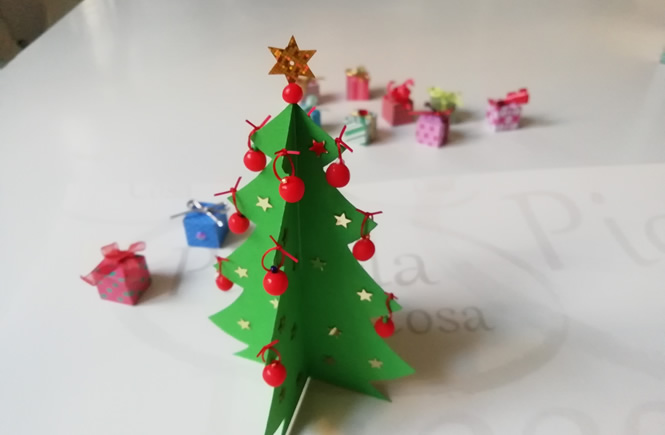 arbol de navidad de cartulina, con bolitas de plastilina en las puntas y estrellitas brillantes pegadas por el cuerpo del árbol. de fondo se ven pequeños regalos dabajo del árbol