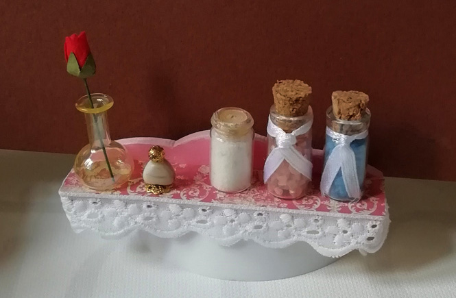 Estantería en miniatura forrada con el papel craft y la puntilla alrededor. Sales de baño, perfume y jarrón de cristal con rosa encima de la estantería.